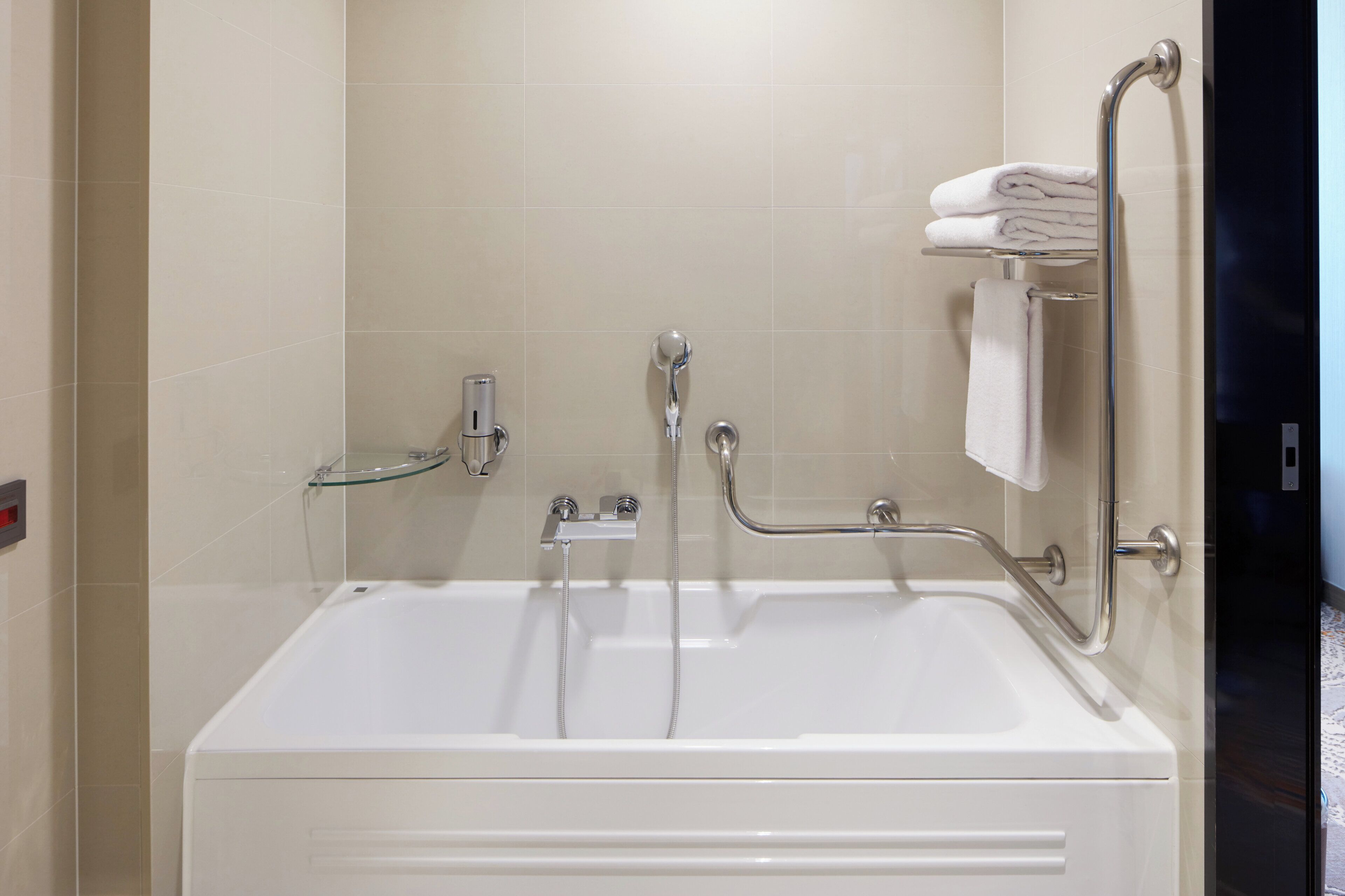A white bathtub with a glass shelf, a handheld showerhead, and a grab bar.