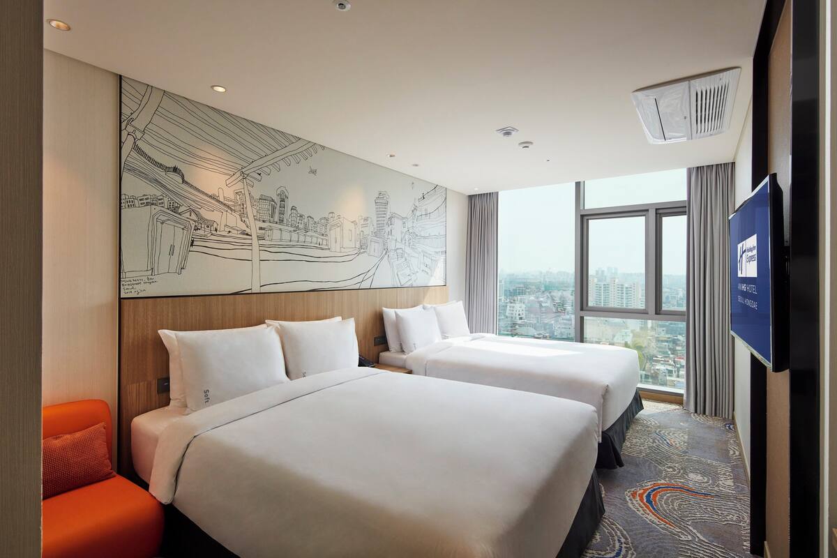 A hotel room with two beds, a large cityscape mural, a television, and a view of the city through large windows.