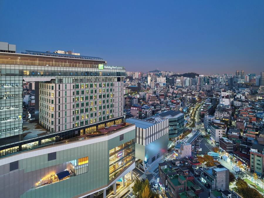 A modern hotel with multiple floors, a prominent Holiday Inn Express sign, and a view of a densely populated cityscape at dusk.