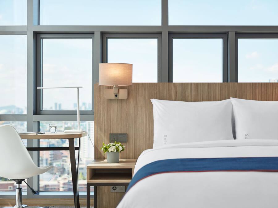 Quarto de hotel com cama, escrivaninha, cadeira, luminária e vista da cidade através de grandes janelas.