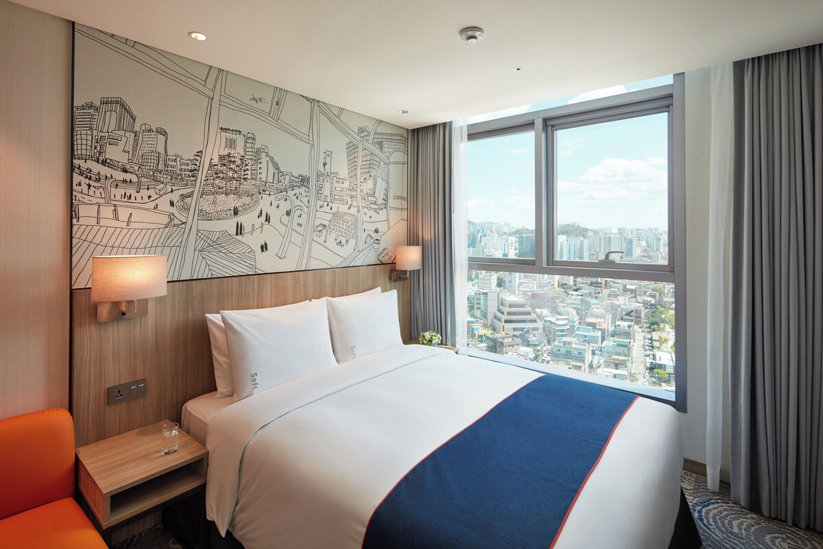 Hotelkamer met groot bed, uitzicht over de stad, een wereldkaart aan de muur en nachtkastje met lamp.