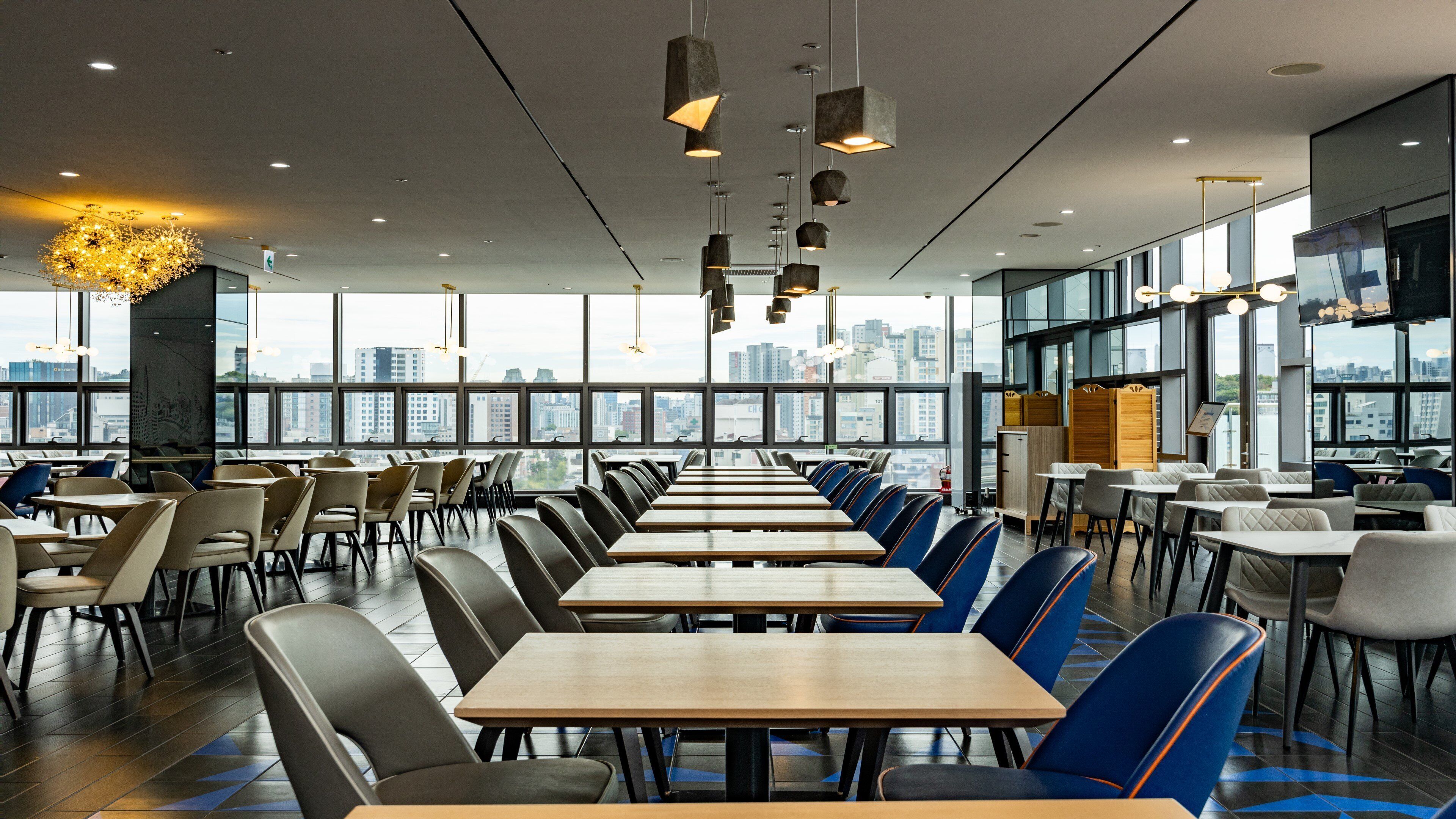 A modern dining area with long tables and chairs, large windows offering city views, and contemporary lighting fixtures.