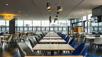 A modern dining area with long tables and chairs, large windows offering city views, and contemporary lighting fixtures.