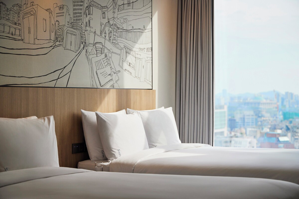 Een hotelkamer met uitzicht op de stad, een bed met witte lakens en een kunstwerk aan de muur dat een stadsgezicht voorstelt.