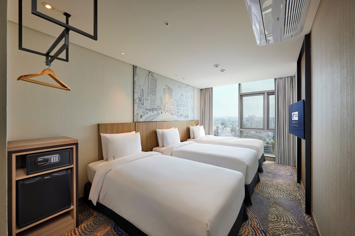 Een hotelkamer met drie bedden, een minibar, een televisie en uitzicht over de stad.