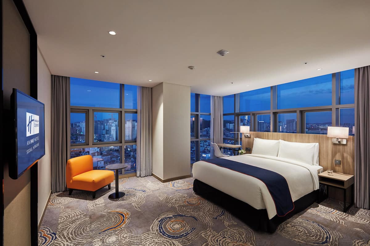 A hotel room with a large bed, a chair, a TV, and a city view through large windows.
