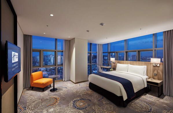 Hotelkamer met een groot bed, een stoel, een televisie en stadszicht door grote ramen.