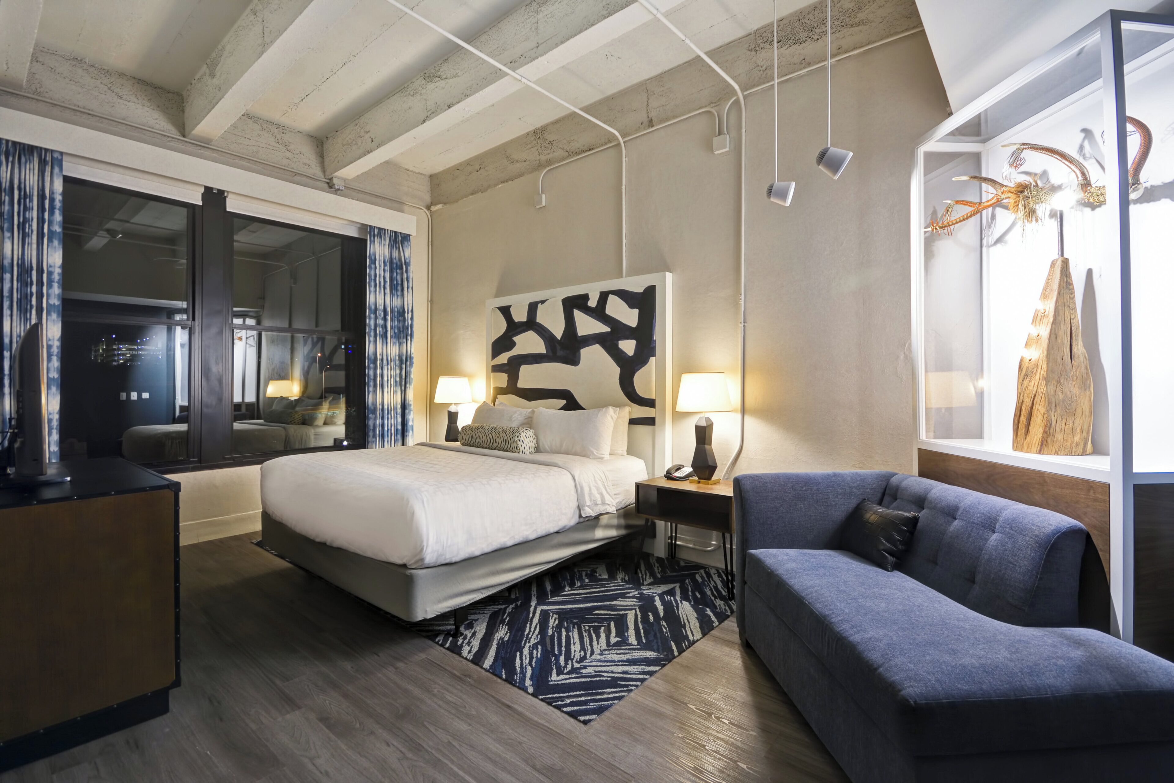 A modern hotel room with a large bed, a sofa, a desk, and a large window.