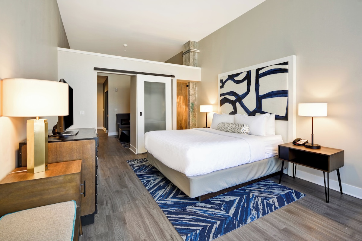 Quarto de hotel com cama, mesas de cabeceira, escrivaninha e uma grande pintura abstrata na parede.
