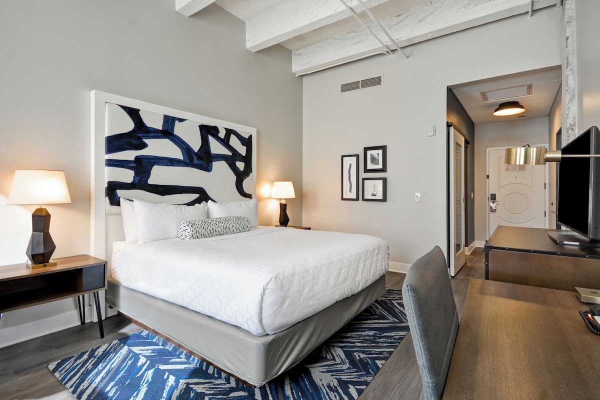 Quarto de hotel moderno com uma cama grande, abajures, escrivaninha e televisão.