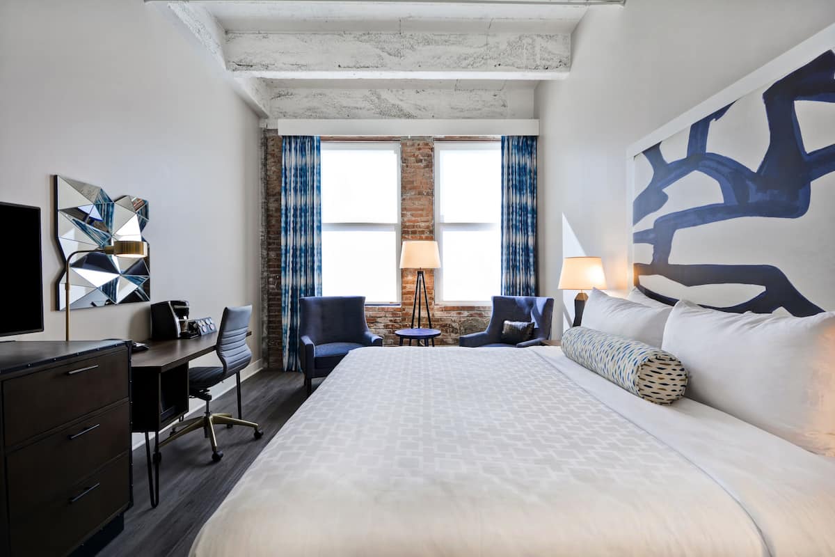 Une chambre d’hôtel avec un grand lit, un bureau avec une chaise, une télévision et une lampe. Il y a deux fenêtres avec des rideaux et un mur en briques derrière.