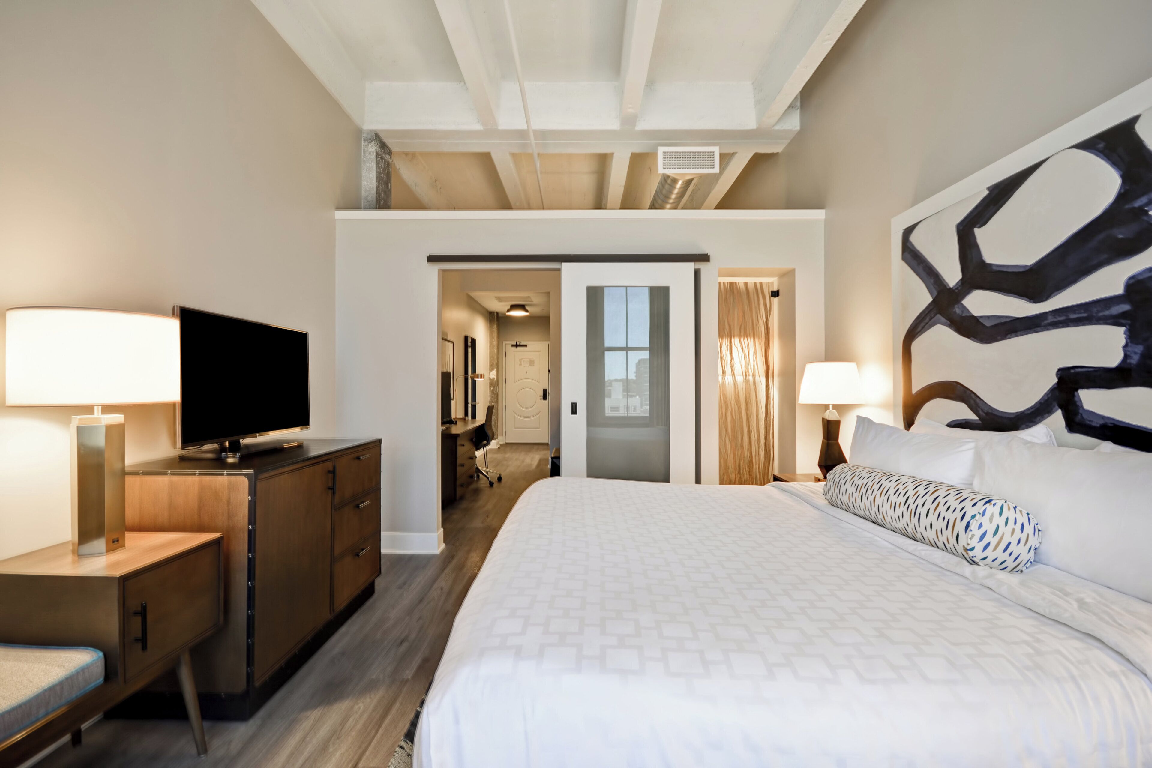 Una habitación de hotel con una cama, un escritorio con televisor, un aparador y un cuadro en la pared.