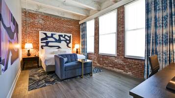 A modern bedroom with exposed brick walls, a large abstract painting, a bed with a blue upholstered headboard, a small side table, and a desk with a chair.