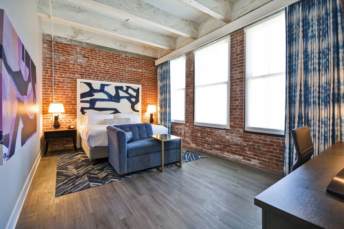 A room with exposed brick walls, a bed with a headboard, two bedside tables with lamps, a blue armchair, a small side table, and a large window with curtains.