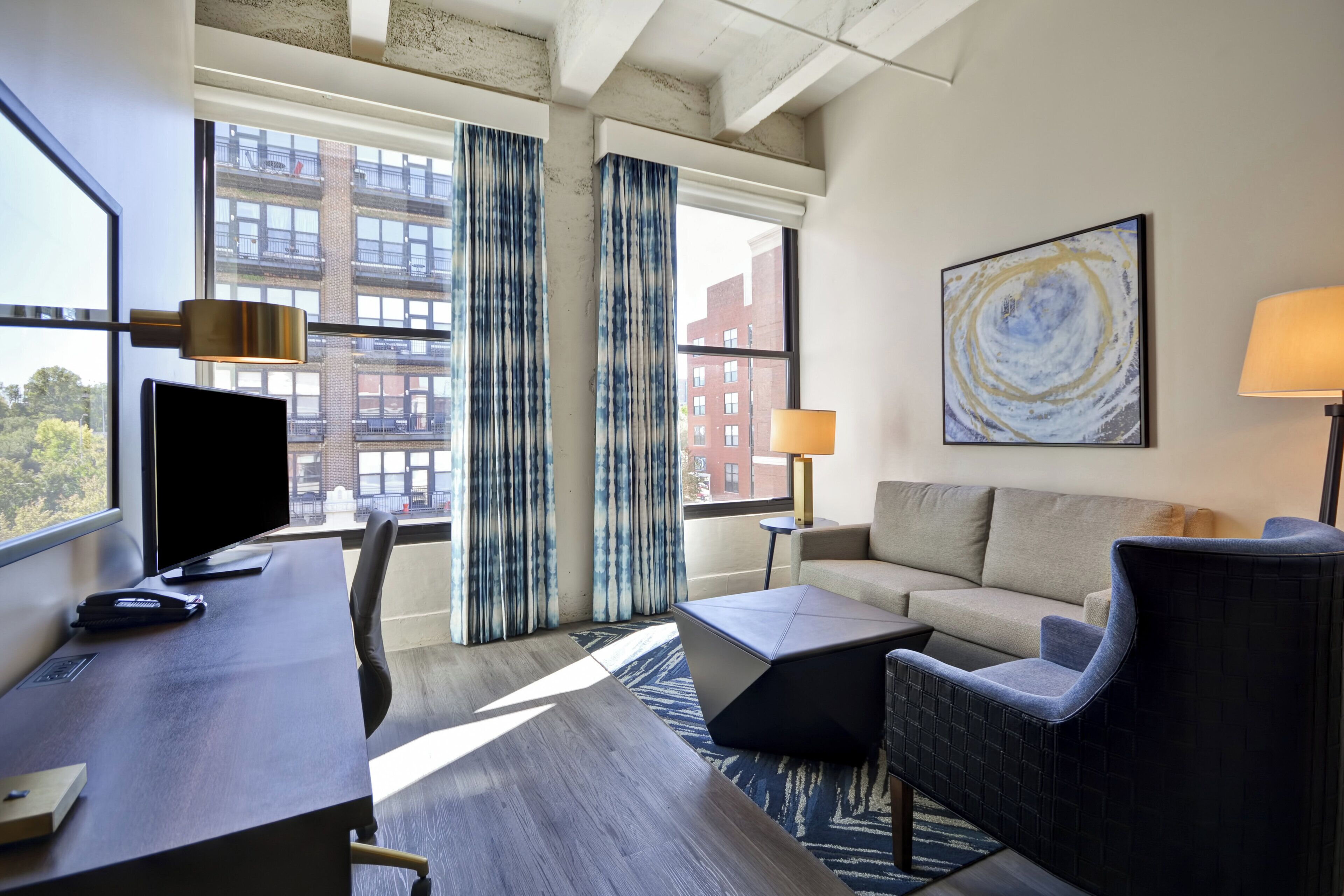 A modern hotel room with a desk, chair, sofa, and a large window with curtains.