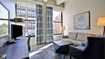 A modern hotel room with a desk, chair, sofa, and a large window with curtains.