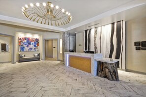 Zona de recepción de hotel con piso de patrón espiga, una lámpara de araña moderna, un mostrador de recepción con base de madera y un cuadro abstracto grande en la pared.