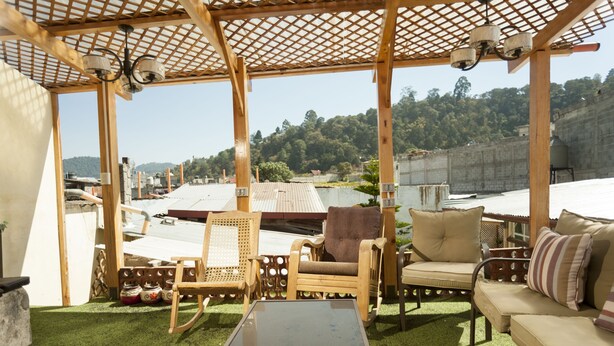 A covered outdoor seating area with wooden furniture and a glass table, overlooking a hilly landscape. - Hotel Vintage Casa las Flores (Quetzaltenango)