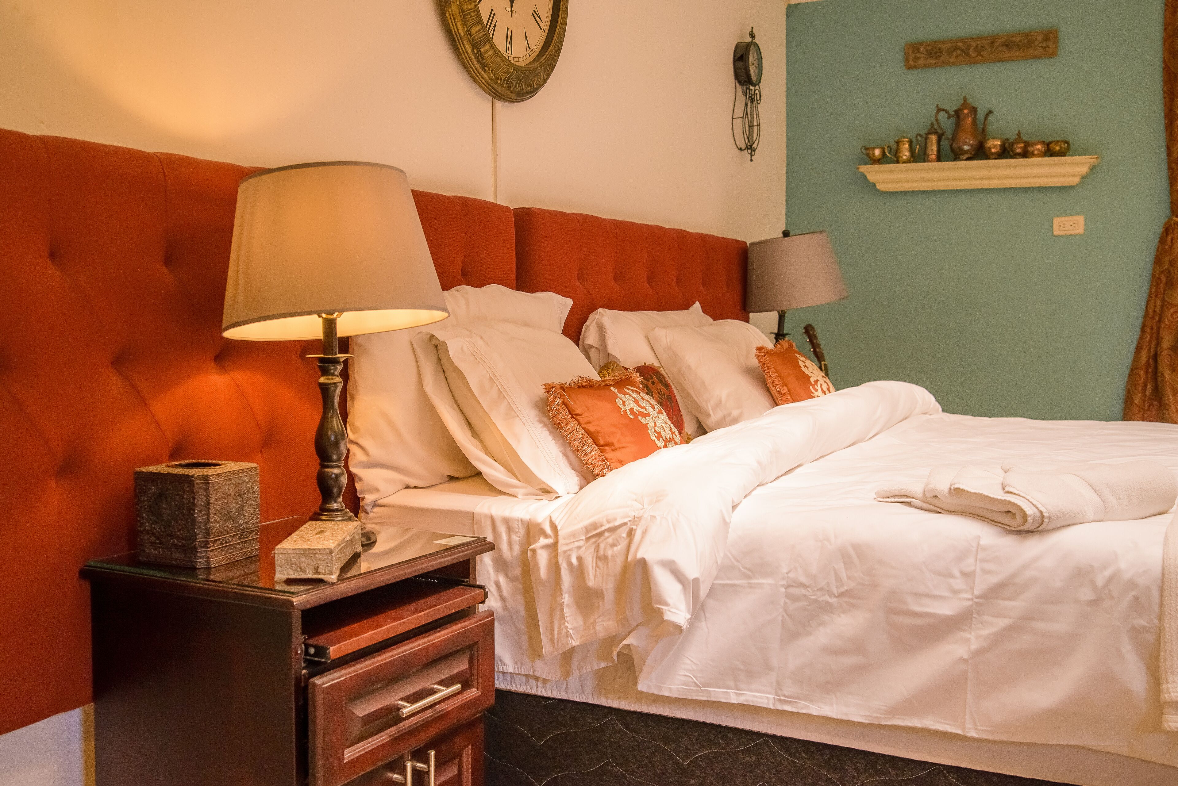 A neatly made bed with white linens, a wooden nightstand with a lamp, and a clock on the wall.