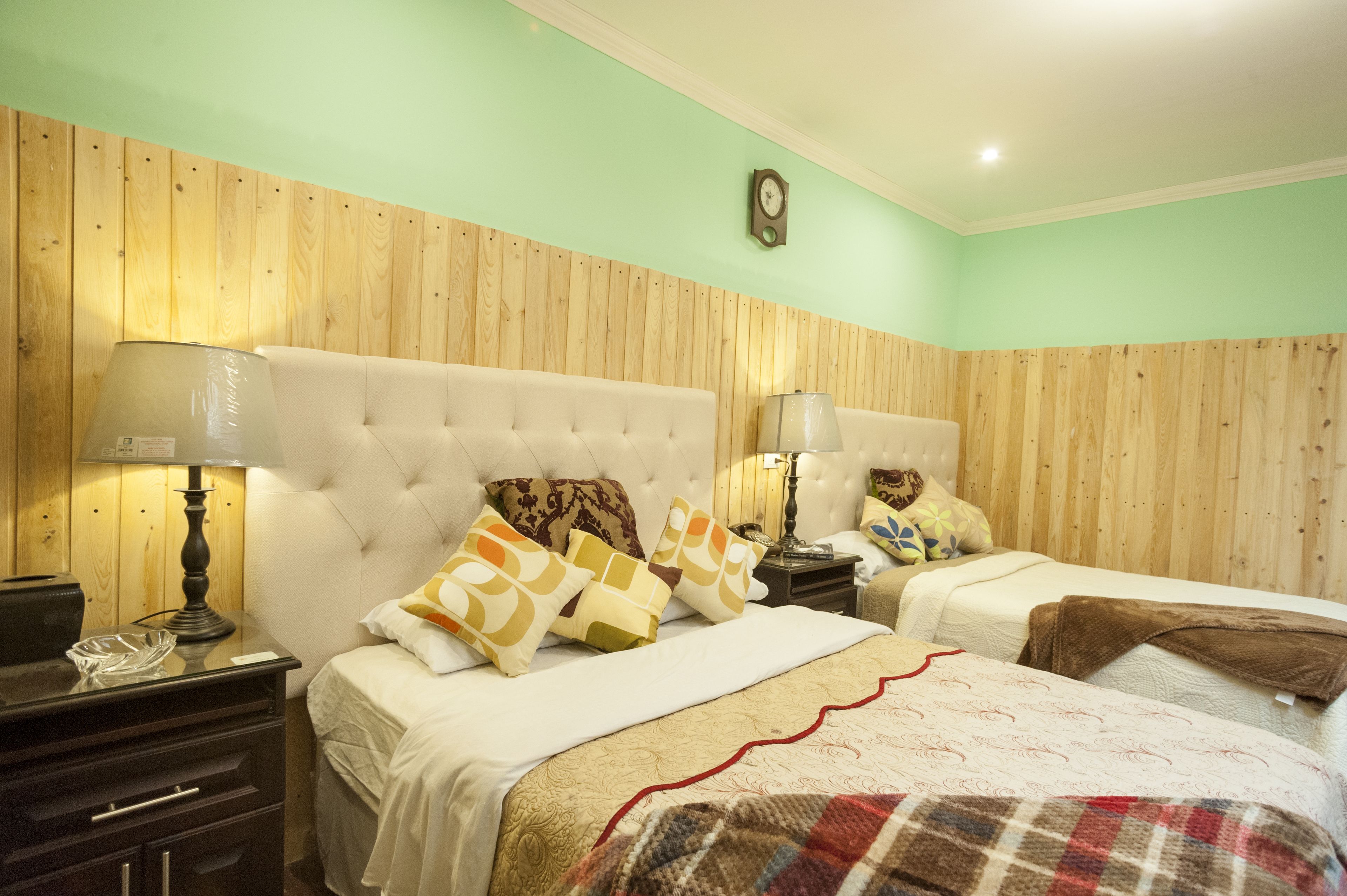 Une chambre avec deux lits, des lambris en bois aux murs, une horloge et une table de chevet avec une lampe.