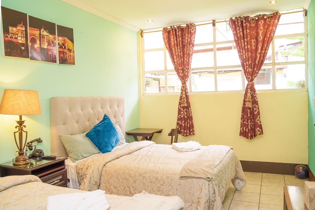Una habitación de hotel con una cama grande, una mesita de noche con lámpara, una pequeña mesa y una ventana con cortinas.