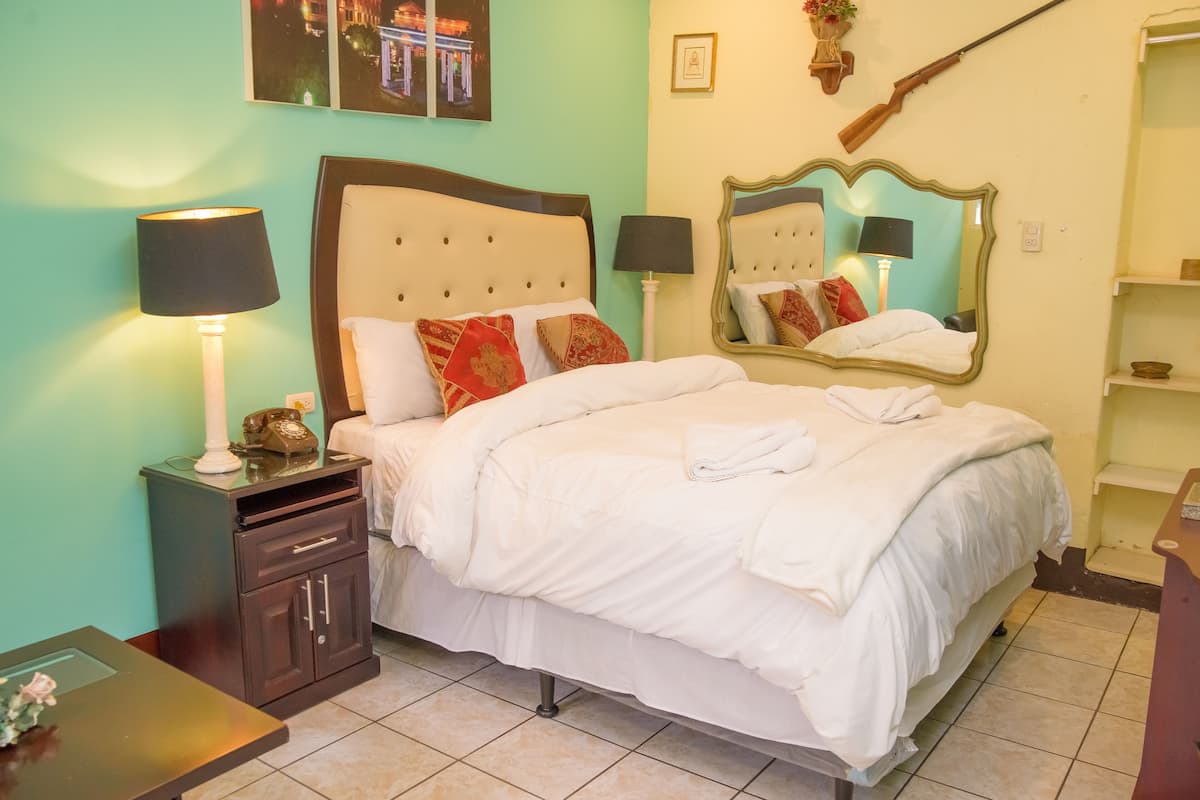 Una cama bien hecha con ropa blanca y almohadas rojas, una mesita de noche con una lámpara, un buró con un teléfono y un espejo en la pared.