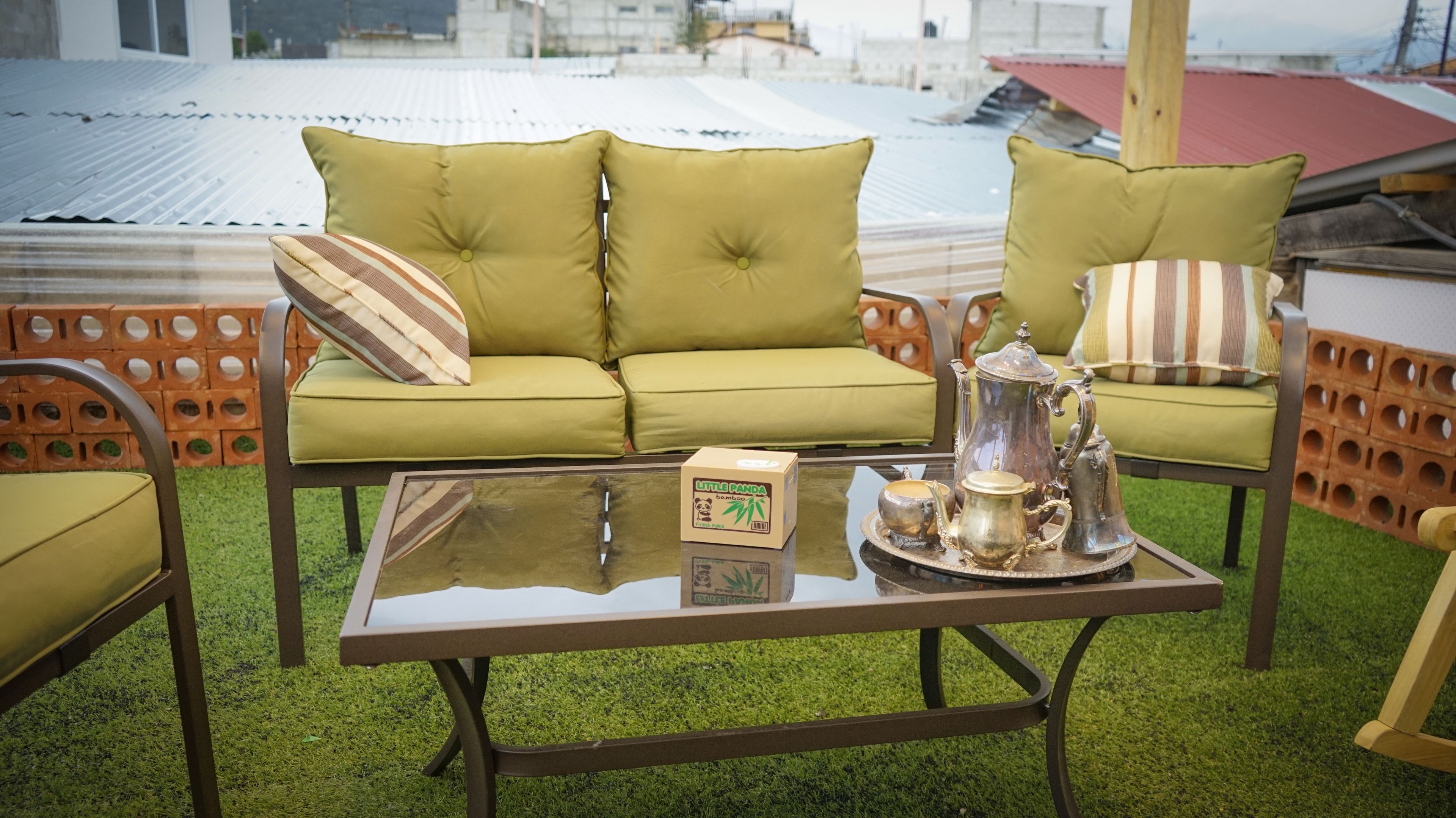 A green sofa with striped cushions, a glass coffee table with a tea set, and a brick wall in the background.