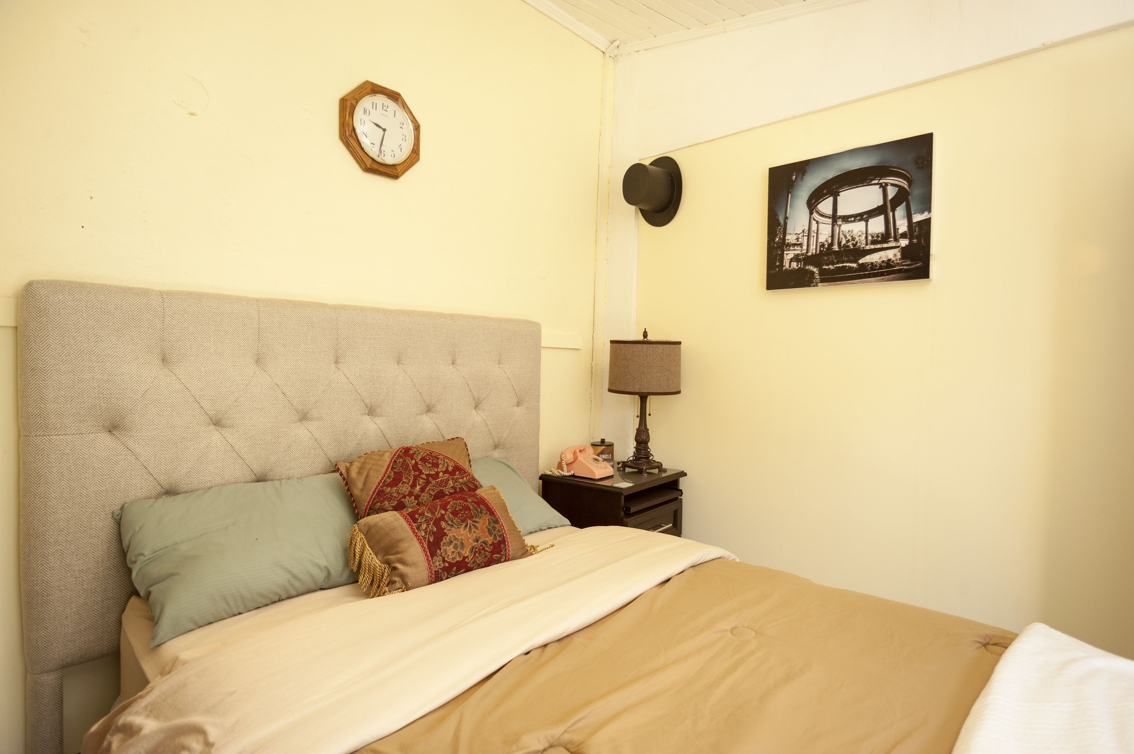 A bedroom with a tufted headboard, a bed with multiple pillows, a bedside table with a lamp, a clock, and a framed picture on the wall.