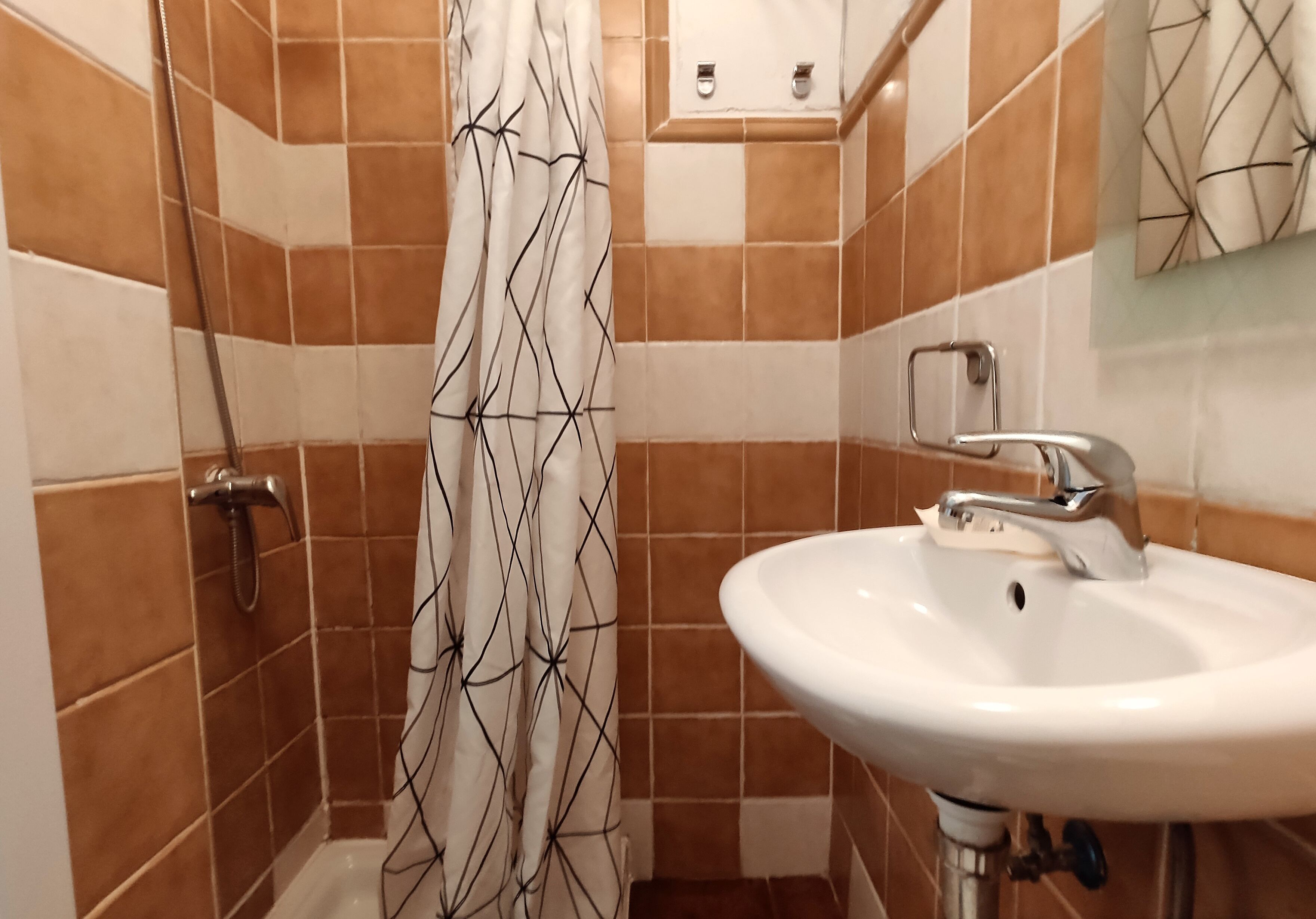 Un baño con azulejos marrones y blancos, una ducha con cortina estampada y un lavamanos blanco con grifo.