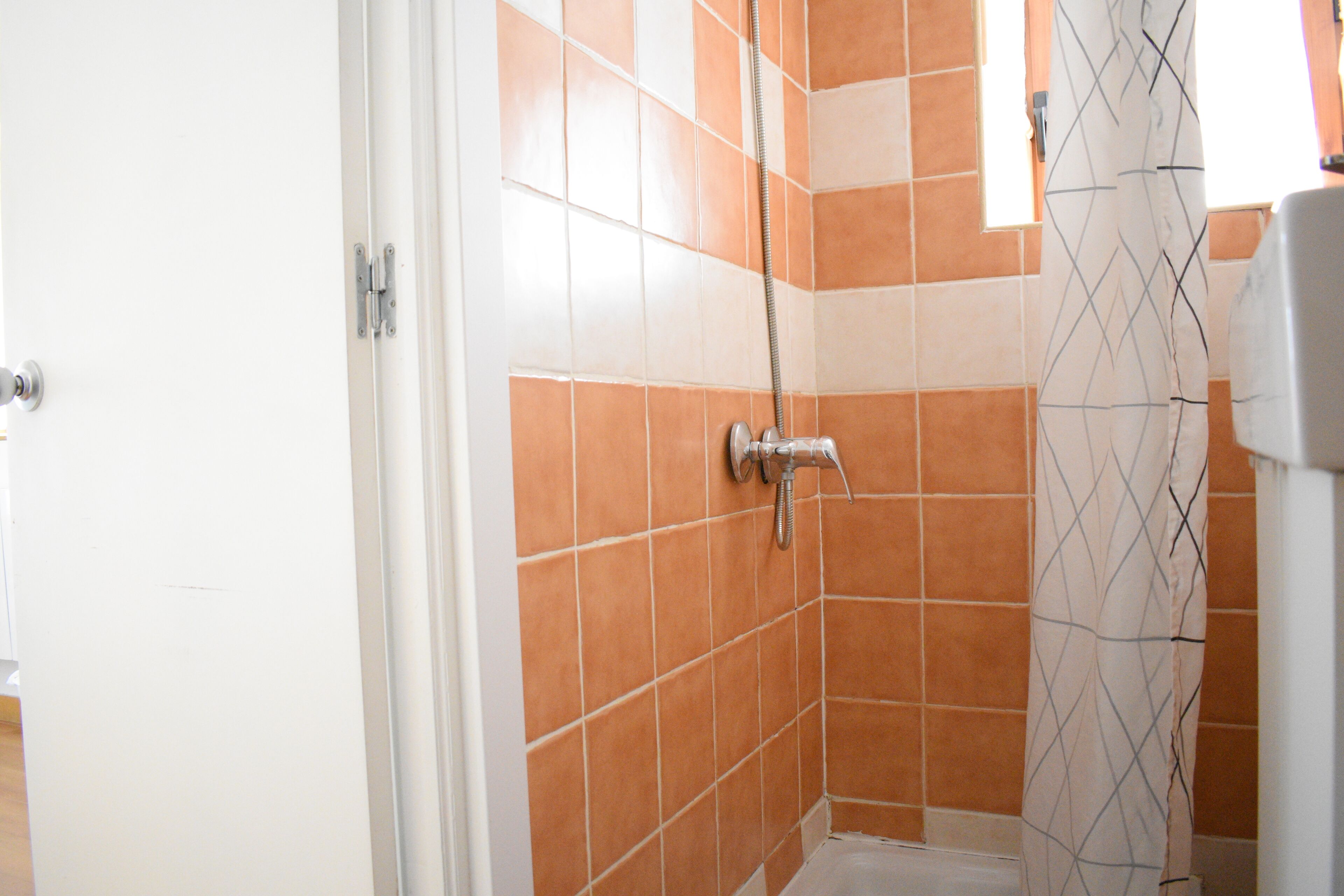 A bathroom with orange and white tiles, a shower with a handheld showerhead, and a white door.