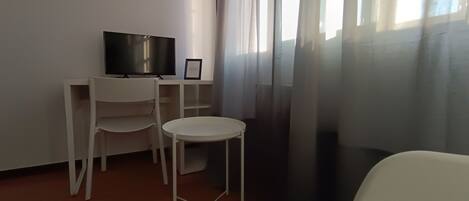 Una habitación pequeña y moderna con un escritorio, una silla, un televisor y una ventana con cortinas de gasa.