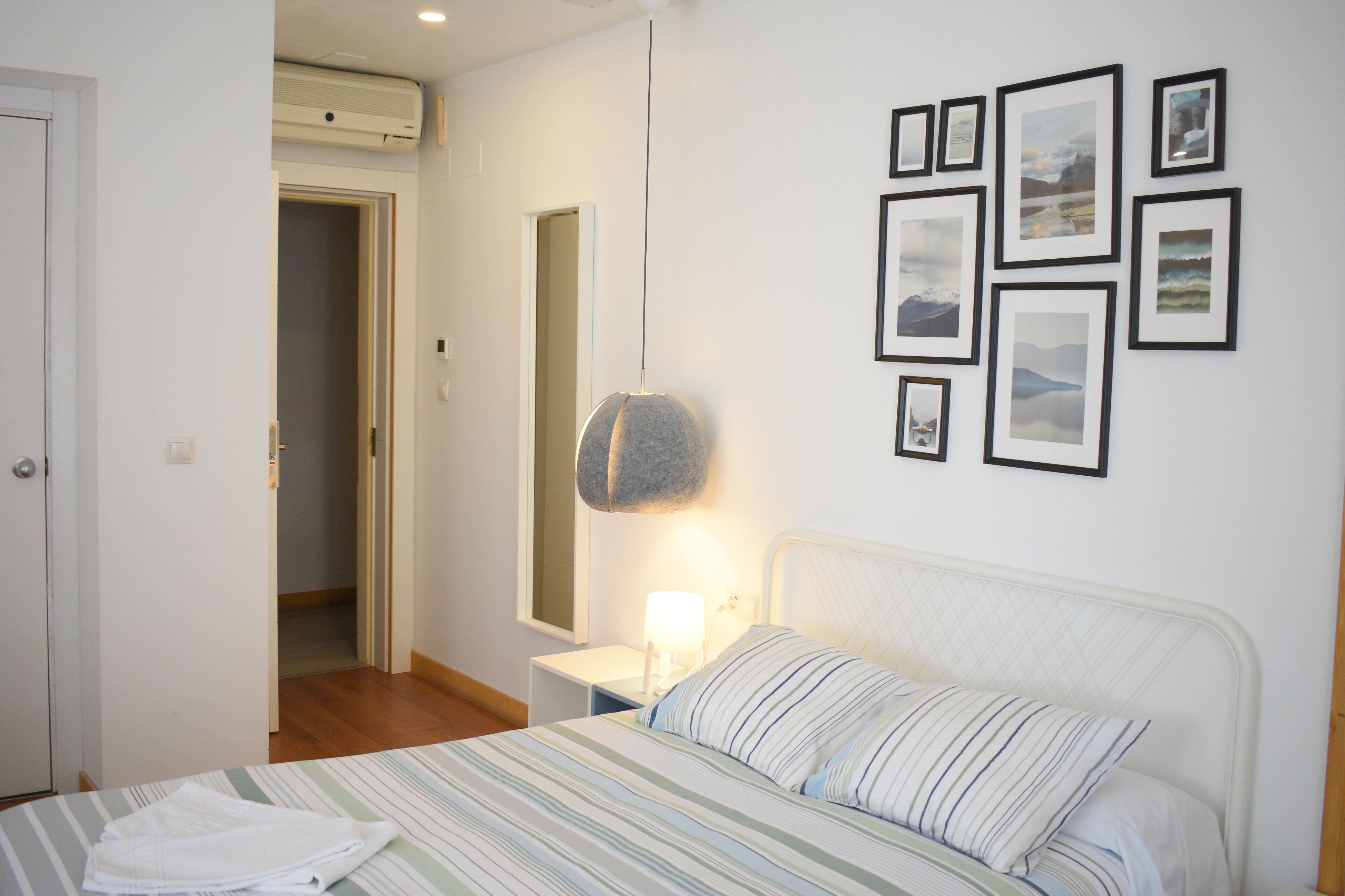 A bedroom with a bed, a bedside table with a lamp, a wall-mounted air conditioner, and framed pictures on the wall.