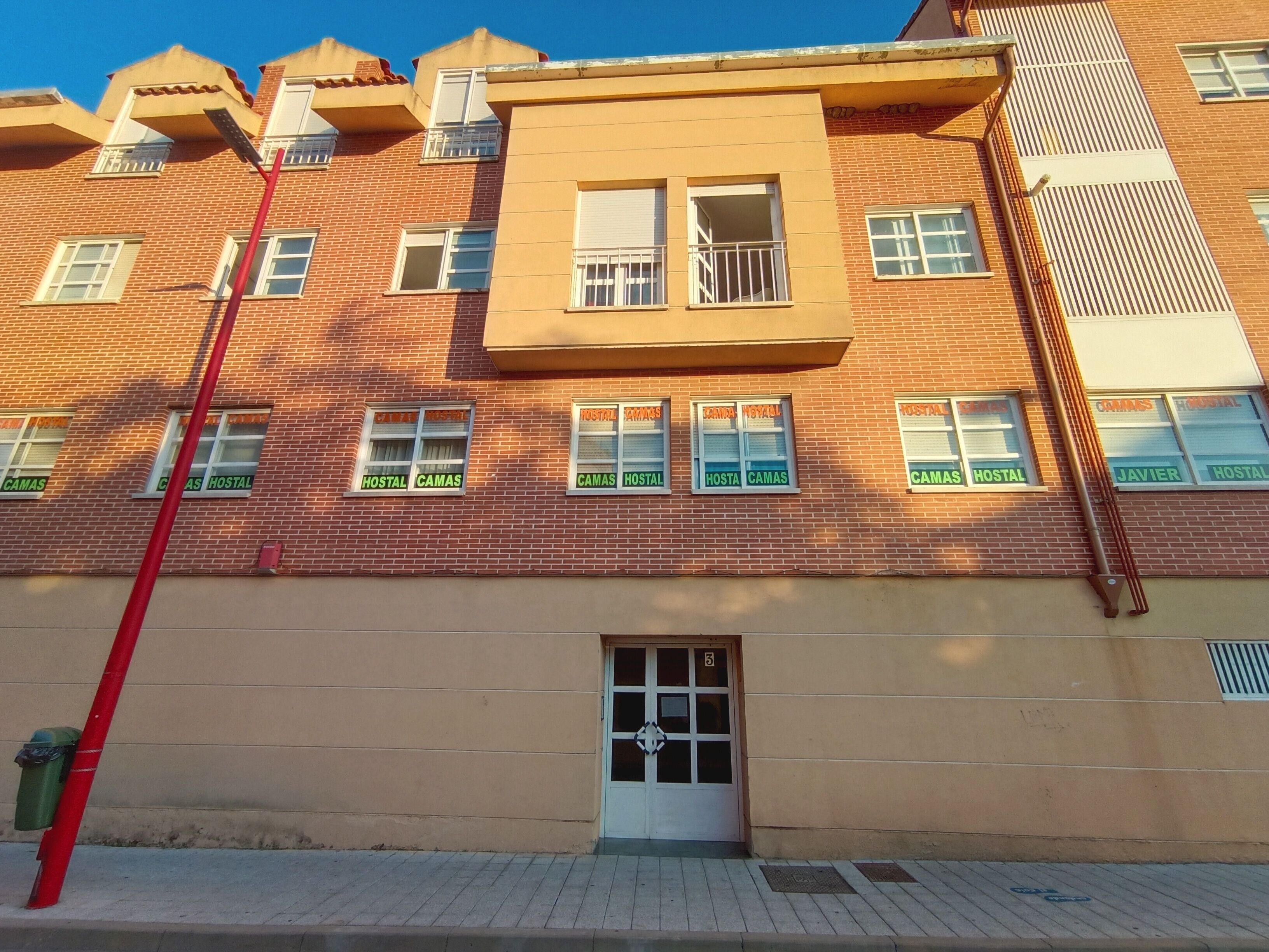 Un edificio de varios pisos con fachada color beige y detalles en ladrillo rojo. Tiene múltiples ventanas con letreros que indican 'Camas Hostal'.