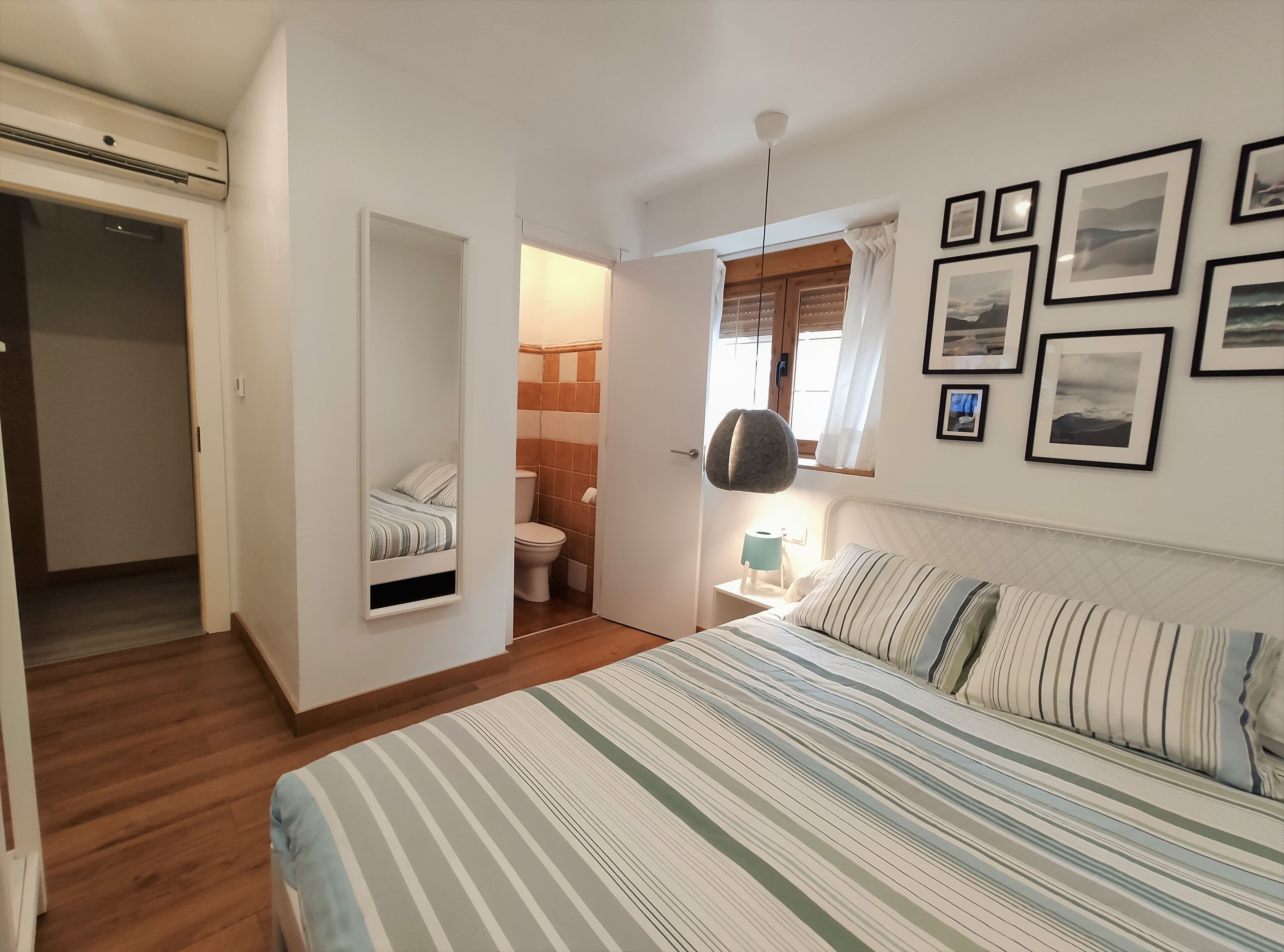 Un dormitorio con cama, mesita de noche, una silla colgante, ventana con cortinas y baño con inodoro y lavabo.