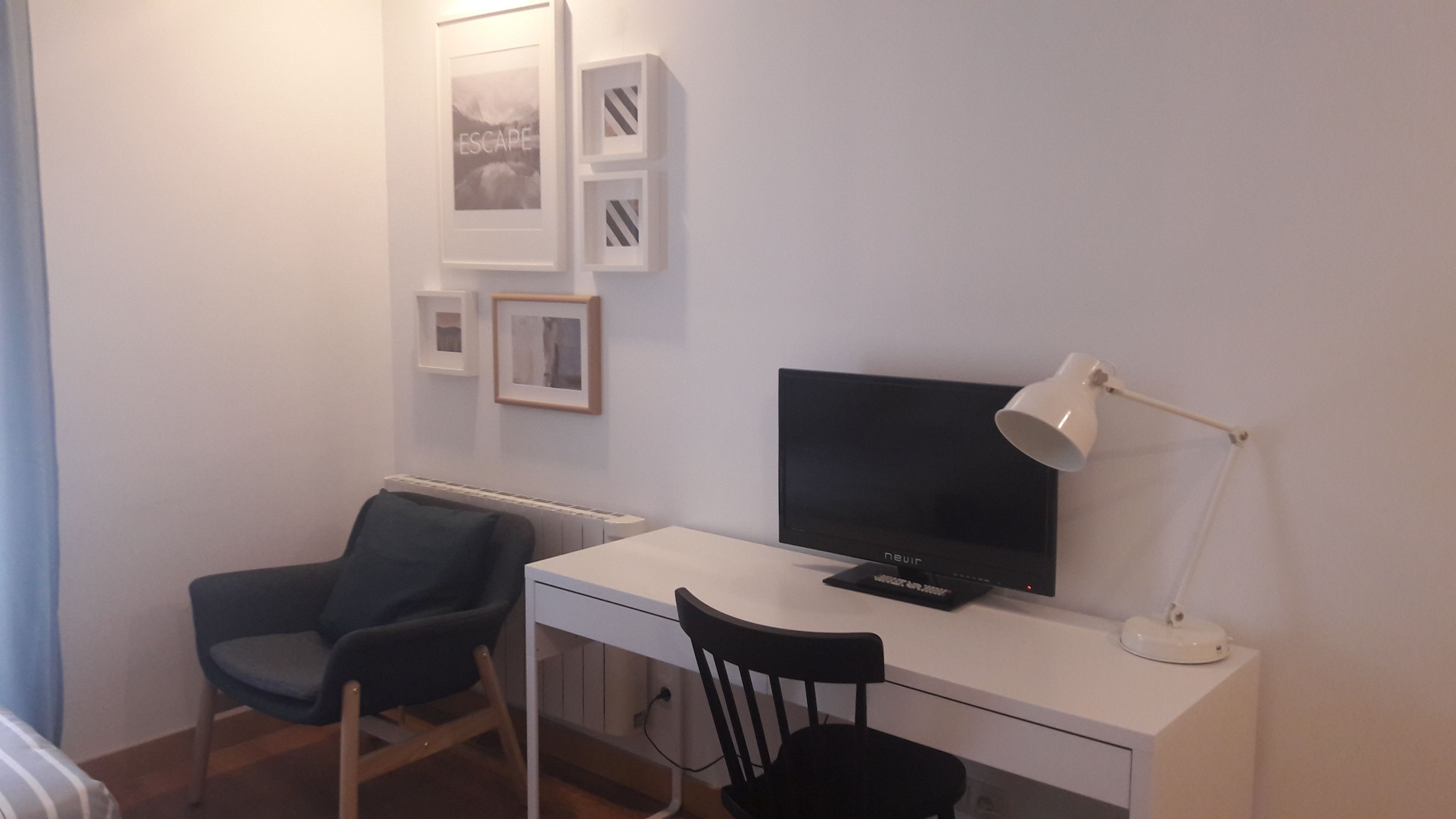 Ett litet, modernt rum med ett vitt skrivbord, en svart stol, en tv och en lampa.