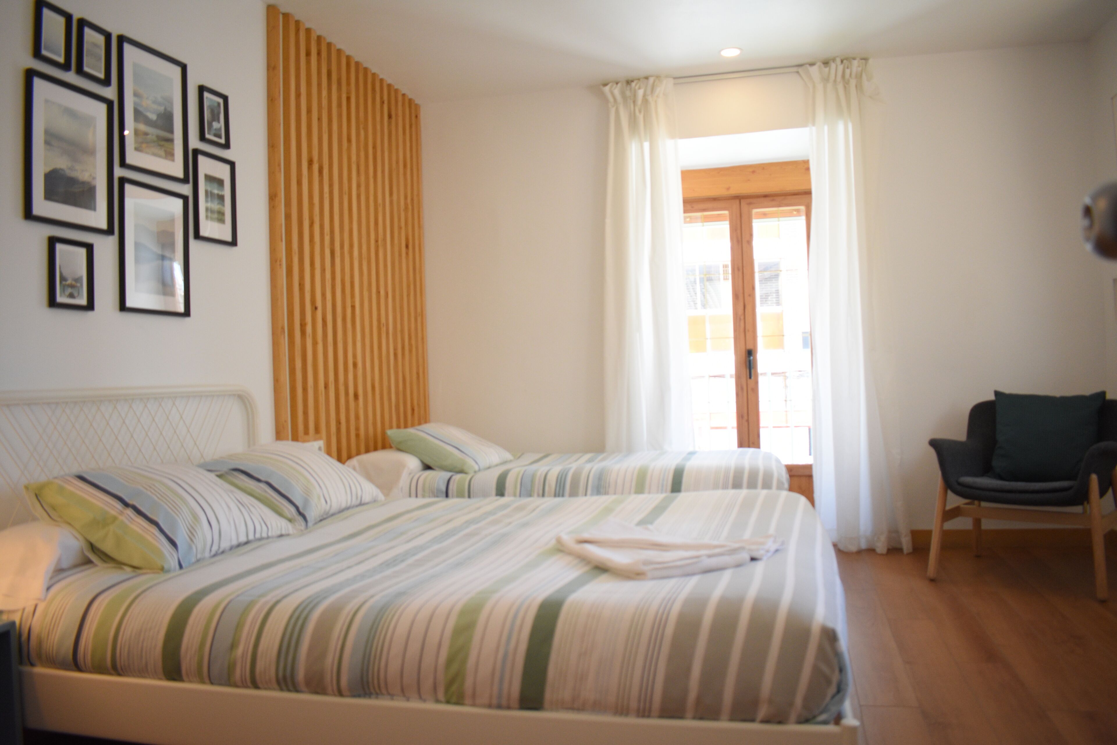 A hotel room with two beds, a wooden panel wall, framed pictures, a chair, and a window with curtains.