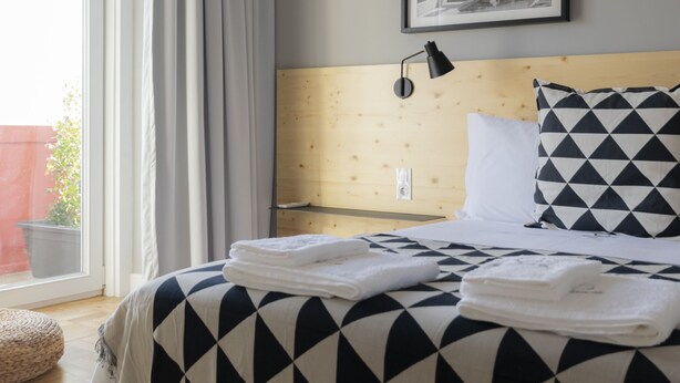 Ein ordentlich bezogenes Bett mit einer Tagesdecke mit geometrischem Muster, ein Kopfteil aus Holzpaneelen, eine Wandlampe und ein gerahmtes Bild darüber.
