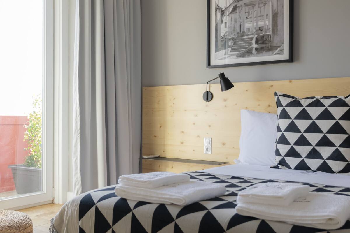 Una cama bien hecha con una manta de estampado geométrico, un cabecero con panel de madera, una lámpara de pared y un cuadro enmarcado sobre la cama.