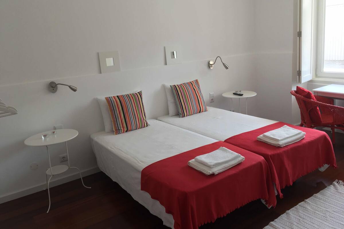 Una habitación de hotel con dos camas, una mesita redonda, una silla roja y una ventana.