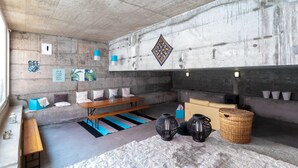 Ein Zimmer mit Betondecke, einer Holzbank, einer Couch und einem kleinen Tisch. Auf dem Boden stehen dekorative Vasen und ein gewebter Korb.