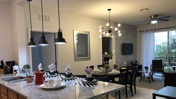 A modern kitchen with granite countertops, hanging pendant lights, and a dining area with a large window offering a view of the outdoors.