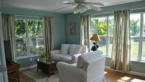 A living room with a sofa, a glass coffee table, and a lamp. There are large windows with curtains, a ceiling fan, and a view of greenery outside. - Tropical Getaway!!!! (Jensen Beach)