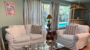 A living room with two white sofas, a glass coffee table, and a view of a swimming pool through large windows. - Tropical Getaway!!!! (Jensen Beach)