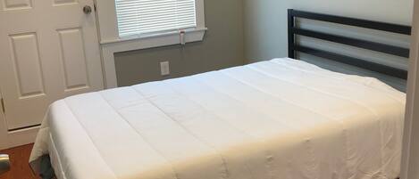 A bedroom with a bed, wooden flooring, a white door, and a window with blinds.