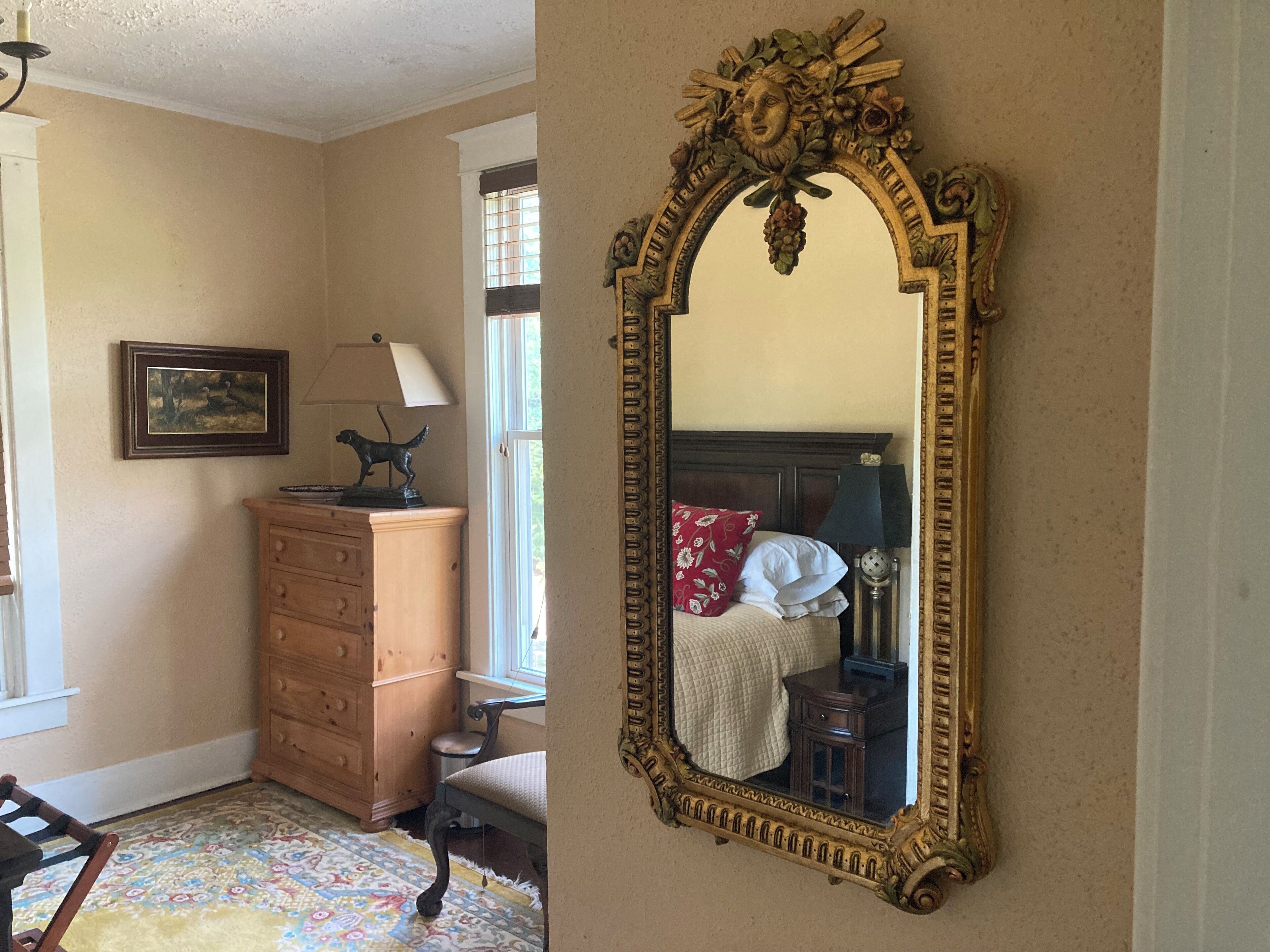Un dormitorio con un espejo grande y decorativo, un tocador de madera, una silla, una cama con almohada roja y una ventana con persianas.