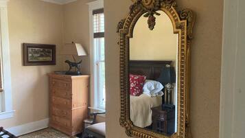 Un dormitorio con un espejo grande y decorativo, un tocador de madera, una silla, una cama con almohada roja y una ventana con persianas.