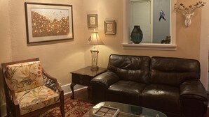 A living room with a leather sofa, a floral armchair, a glass coffee table, a side table with a lamp, and a framed picture on the wall. - Pegasus Lair Upper Floor Apt.NoPetFee, NoChores.Short Walk to Satellite Venue! (Memphis)