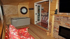 A cozy cabin interior with a wooden floor, a red patterned sofa, a wooden bathtub, a fireplace, and a large window with a bed visible through it. - Gatlinburg Cabin Free Tickets, Heart Whirlpool, Hot Tub, King Bed, Fast Internet (Gatlinburg)