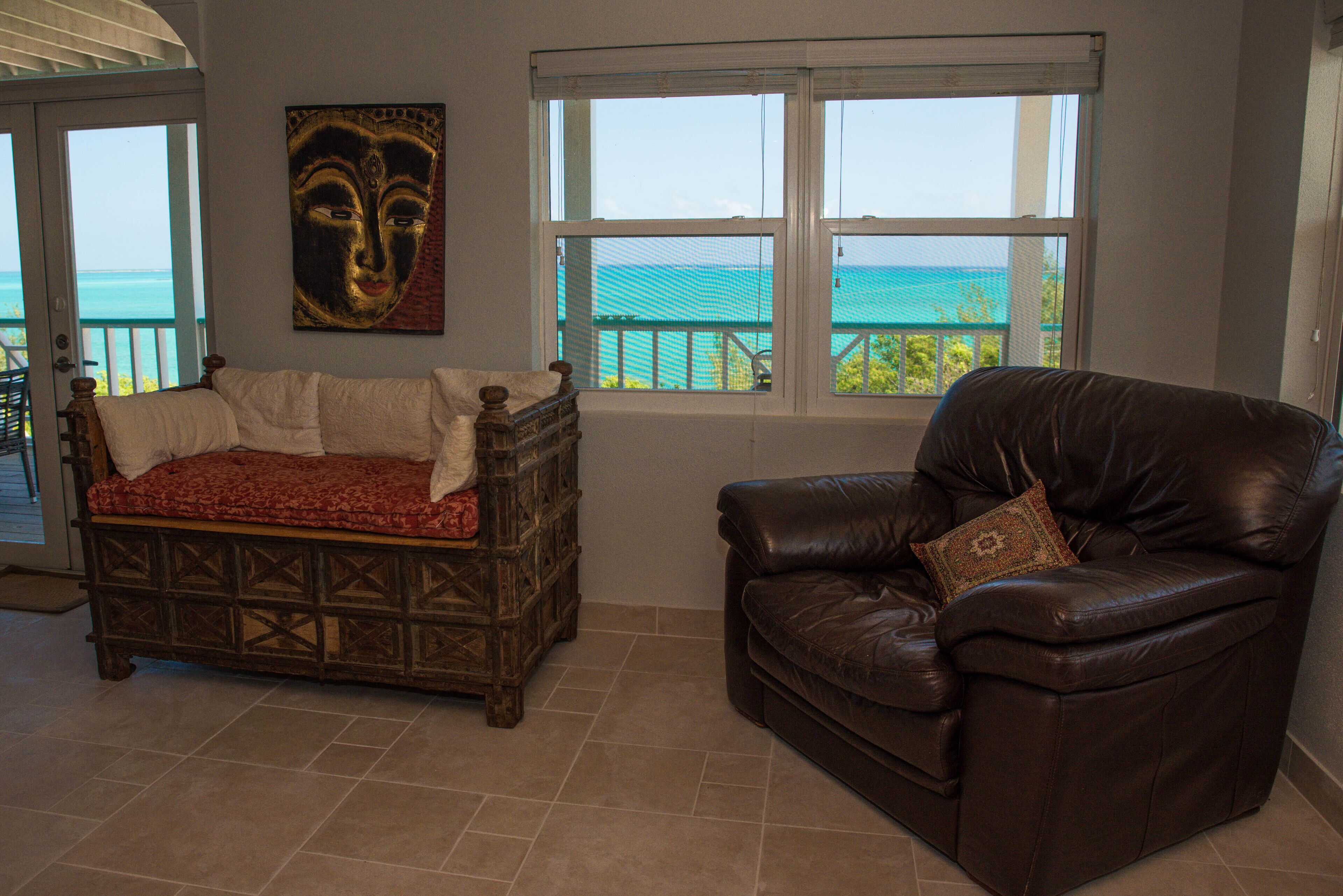 A room with a wooden bench and a leather chair, a painting on the wall, and a view of the ocean through windows.