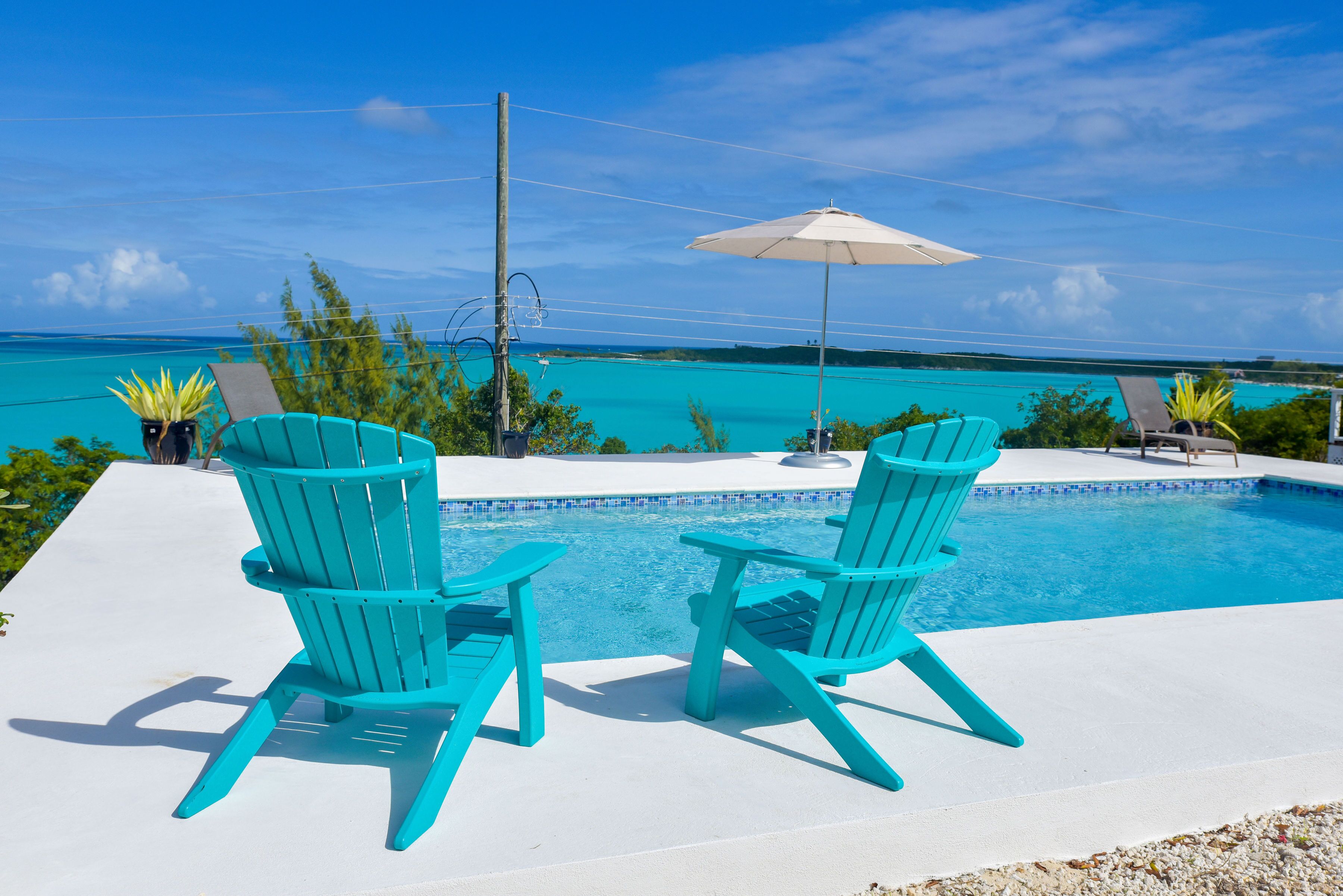 A pool area with two turquoise Adirondack chairs and an umbrella, surrounded by a clear blue sky and ocean view.